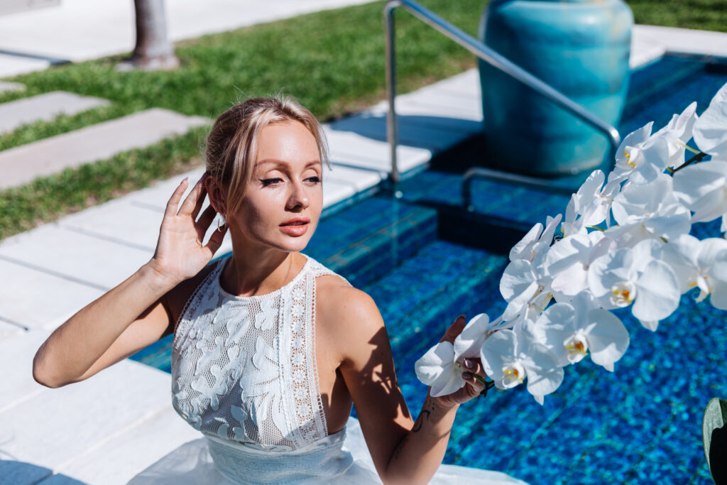 outdoor-portrait-woman-white-wedding-dress-sitting-near-blue-swimming-pool-with-flowers-orchid